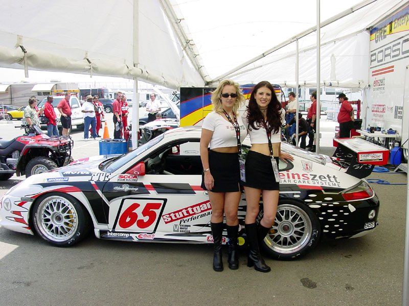 Two women posing in front of a white and black einszett race car with red and white decals, number 65, under a white event tent.