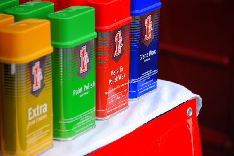Four einszett metallic paint polish containers in yellow, green, red, and blue, labeled with the same red-and-black shield einszett logo, arranged on a white cloth next to a red surface.