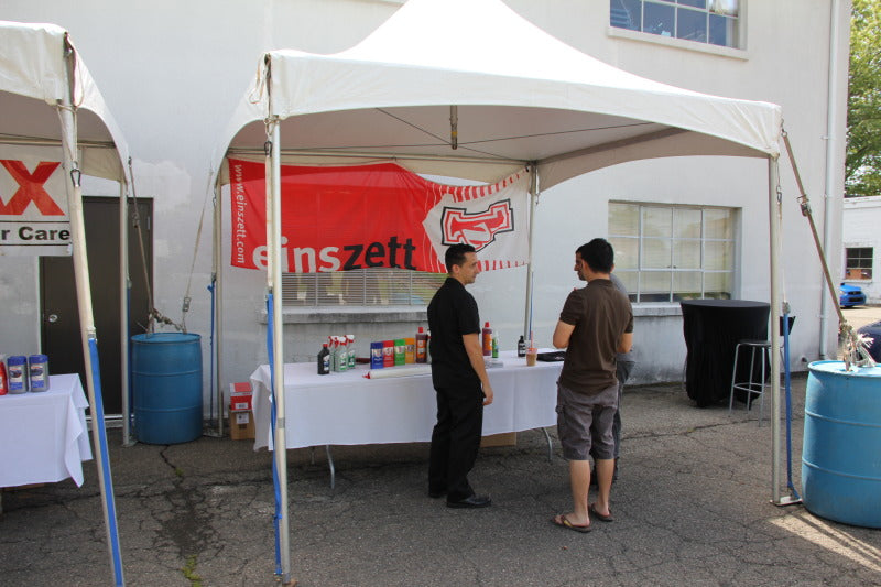 A white pop-up canopy tent set up outdoors in front of a white building, with a red and white banner hanging behind it. Two men stand under the tent at a table with bottles and other items on display.