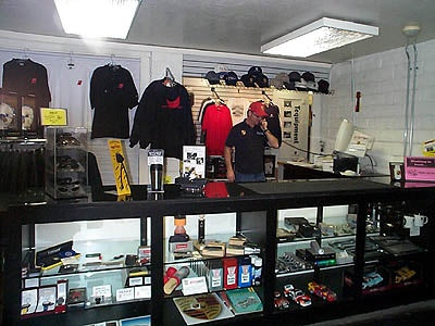 A retail display counter at Newport Beach Porsche (2001) filled with small merchandise, with several black and red shirts hanging on the wall behind, a person standing in the background, and fluorescent ceiling lights overhead.