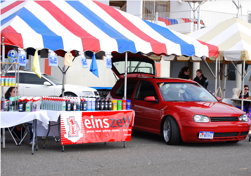 A red Volkswagen Golf parked outdoors under a red, white, and blue striped canopy, with its trunk open. A market or fair booth with tables, einszett bottles, and a banner reading 'einszett' is set up beside it.