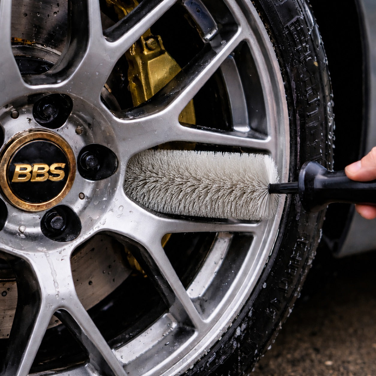A close-up of a hand using a wheel barrel brush with soft bristles to clean a silver alloy car wheel with a visible BBS logo.