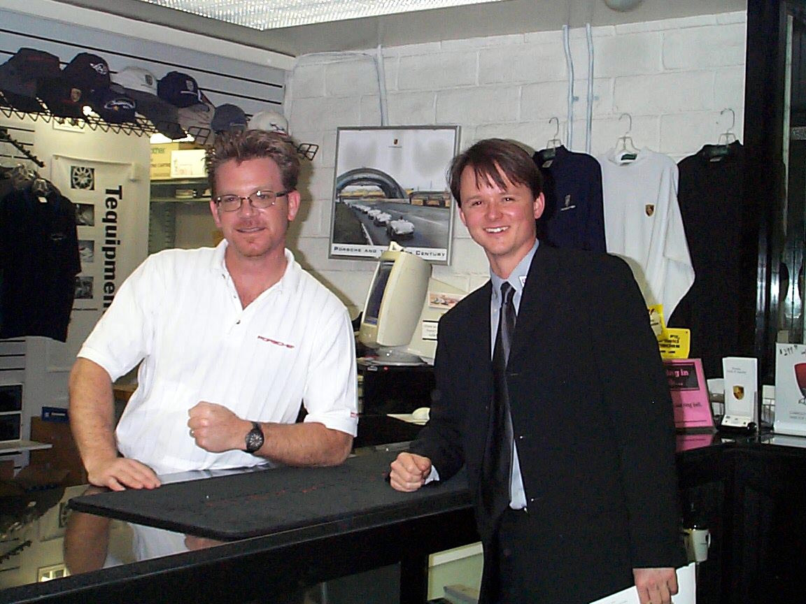 Two men standing behind a counter in a retail store. The man on the left wears a white polo shirt, and the man on the right wears a dark suit jacket, light blue shirt, and dark tie.