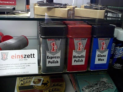 Three Einszett wax containers on a store shelf at Newport Beach Porsche (2001) Express Polish in gray, Metallic Polish in red, and Glanz Wax in blue, with a white Einszett sign beside them.