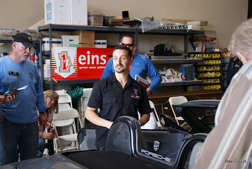 A man in a black einszett work shirt stands behind a black Porsche car in a workshop, with other people and shelves visible in the background. A red 'einszett German vehicle parts' sign is on the wall behind him.