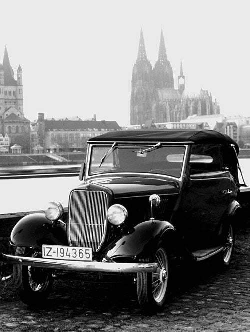 A vintage black convertible car parked outdoors in front of a cityscape with a large cathedral spire in the background, shown in black-and-white.
